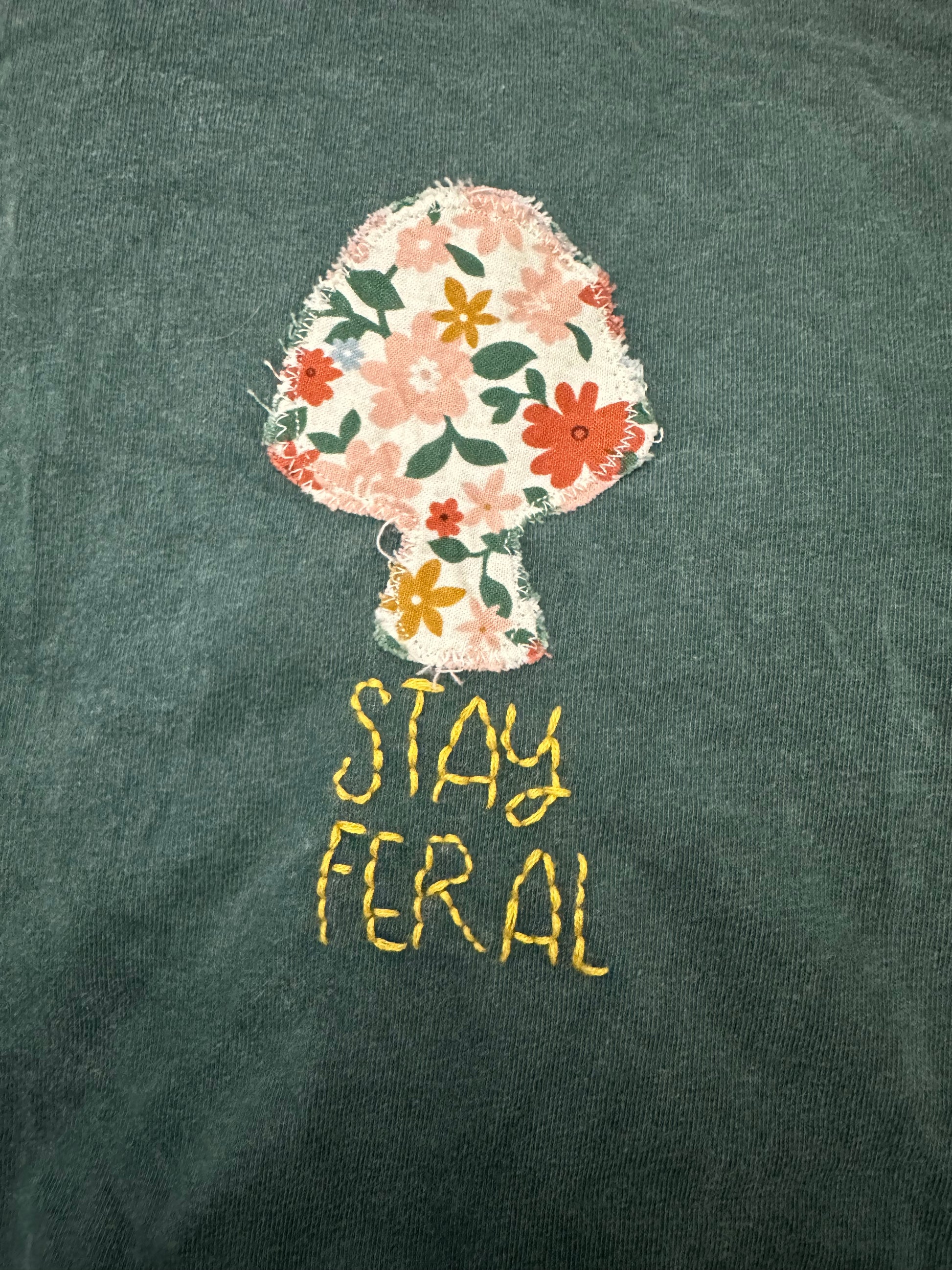 The image shows a floral mushroom-shaped design embroidered on a dark green fabric, with the text "STAY FERAL" written below it in yellow thread.